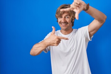 Poster - Middle age man standing over blue background smiling making frame with hands and fingers with happy face. creativity and photography concept.