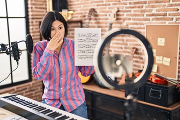 Poster - Young asian woman doing online music tutorial showing music sheet covering mouth with hand, shocked and afraid for mistake. surprised expression