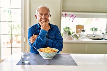 Canvas Print - Senior man with grey hair eating pasta spaghetti at home looking confident at the camera smiling with crossed arms and hand raised on chin. thinking positive.