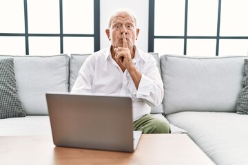 Canvas Print - Senior man using laptop at home sitting on the sofa asking to be quiet with finger on lips. silence and secret concept.