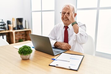 Canvas Print - Senior man working at the office using computer laptop doing ok gesture with hand smiling, eye looking through fingers with happy face.