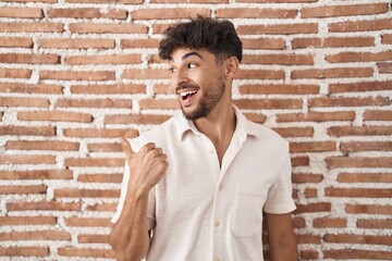 Poster - Arab man with beard standing over bricks wall background smiling with happy face looking and pointing to the side with thumb up.