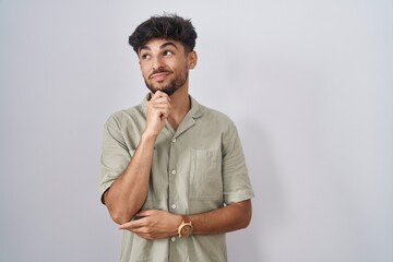 Poster - Arab man with beard standing over white background with hand on chin thinking about question, pensive expression. smiling with thoughtful face. doubt concept.