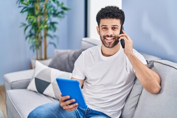 Wall Mural - Young arab man talking on the smartphone using touchpad sitting on sofa at home