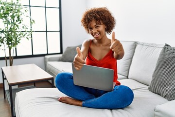 Sticker - Young african american woman sitting on the sofa at home using laptop approving doing positive gesture with hand, thumbs up smiling and happy for success. winner gesture.