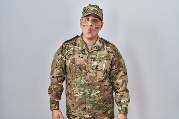 Hispanic young man wearing camouflage army uniform making fish face with lips, crazy and comical gesture. funny expression.