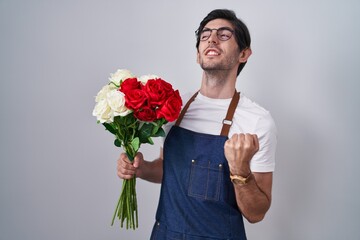 Poster - Young hispanic man holding bouquet of white and red roses very happy and excited doing winner gesture with arms raised, smiling and screaming for success. celebration concept.