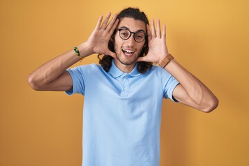 Poster - Young hispanic man standing over yellow background smiling cheerful playing peek a boo with hands showing face. surprised and exited