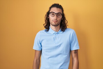 Poster - Young hispanic man standing over yellow background relaxed with serious expression on face. simple and natural looking at the camera.