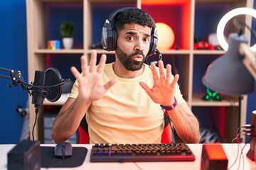 Poster - Hispanic man with beard playing video games with headphones afraid and terrified with fear expression stop gesture with hands, shouting in shock. panic concept.