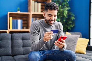 Sticker - Young hispanic man using smartphone and credit card sitting on sofa at home