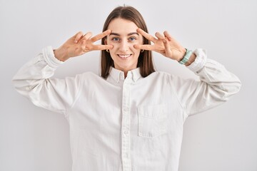 Poster - Young caucasian woman standing over isolated background doing peace symbol with fingers over face, smiling cheerful showing victory