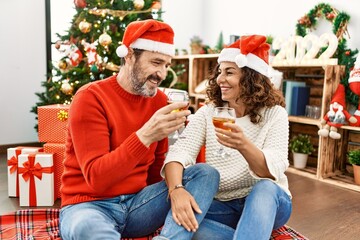 Poster - Middle age hispanic couple wearing christmas hat. Sitting on the floor toasting with champagne at home.