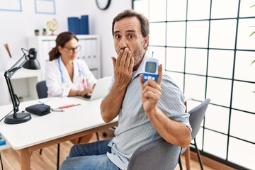 Middle age man at doctor clinic holding glucose meter device covering mouth with hand, shocked and afraid for mistake. surprised expression