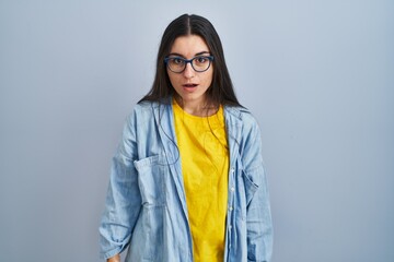 Poster - Young hispanic woman standing over blue background afraid and shocked with surprise and amazed expression, fear and excited face.