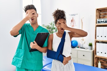 Canvas Print - Physiotherapist working with patient wearing arm on sling at rehabilitation clinic peeking in shock covering face and eyes with hand, looking through fingers with embarrassed expression.