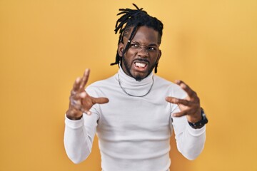 Wall Mural - African man with dreadlocks wearing turtleneck sweater over yellow background shouting frustrated with rage, hands trying to strangle, yelling mad