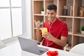 Sticker - Young hispanic man using laptop and doing video call with smartphone pointing thumb up to the side smiling happy with open mouth