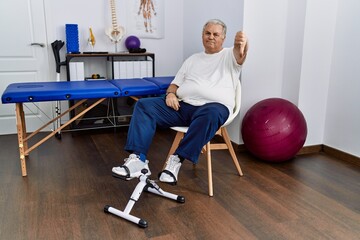 Wall Mural - Senior caucasian man at physiotherapy clinic using pedal exerciser looking unhappy and angry showing rejection and negative with thumbs down gesture. bad expression.