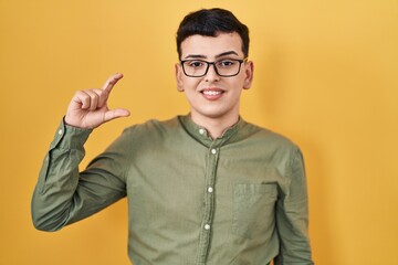 Sticker - Non binary person standing over yellow background smiling and confident gesturing with hand doing small size sign with fingers looking and the camera. measure concept.