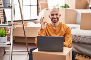 Wall Mural - Young bald man with beard moving to a new house using laptop doing ok sign with fingers, smiling friendly gesturing excellent symbol