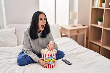 Canvas Print - Hispanic woman eating popcorn watching a movie on the bed angry and mad screaming frustrated and furious, shouting with anger. rage and aggressive concept.