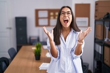 Young Hispanic Woman At The Office Crazy And Mad Shouting And Yelling With Aggressive Expression And Arms Raised. Frustration Concept.