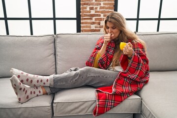 Sticker - Young woman sitting on the sofa drinking a coffee at home feeling unwell and coughing as symptom for cold or bronchitis. health care concept.