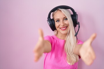 Poster - Caucasian woman listening to music using headphones looking at the camera smiling with open arms for hug. cheerful expression embracing happiness.