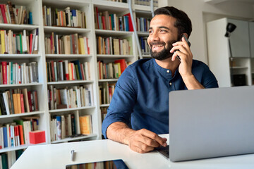 Wall Mural - Happy smiling eastern indian professional business man sitting at work desk talking on mobile phone, ethnic businessman making mobile phone call by cellphone working at home or in office.