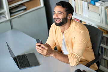 Wall Mural - Happy smiling ethnic indian professional business man manager sitting at work desk wearing earbud using cell phone, laughing eastern businessman having virtual mobile chat video call on smartphone.