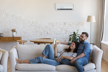 Couple Enjoy Fresh Conditioned Air Inside Cozy Living Room. Carefree Wife Holds Remote Controller Adjust Comfortable Temperature Relaxing On Couch With Husband At Home. Tech For Comfort Life Concept