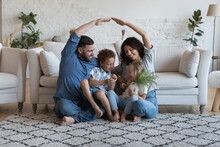 Couple And 5s Kids Sit On Floor In Cozy Living Room, Loving Parents Makes Symbol Of Roof Protecting Their Children, Celebrate First Day At New Own House. Bank Loan For Young Family, Relocation Concept