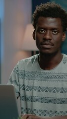 Poster - Vertical video: Close up of african american man working on laptop. Black employee doing remote work on internet while looking at camera and smiling. Business person of african ethnicity using device