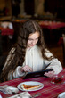 A cute teenage girl sits in a cafe with a gadget in her hands. She has long hair. She is wearing a beige hoodie. In front of her is a plate of pizza. Communication online. Use gadgets. Generation Z.