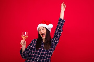 Wall Mural - A smiling young woman in pyjamas wearing a Santa Claus hat holds a glass of wine on a red background. New Year's Eve at home