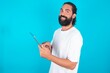 © Roquillo - Photo of happy cheerful smart young bearded man wearing white T-shirt over blue studio background hold tablet browsing internet