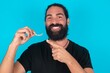 © Roquillo - young bearded man wearing black T-shirt over blue studio background holding an invisible aligner and pointing at it. Dental healthcare and confidence concept.