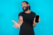 © Roquillo - young bearded man wearing black T-shirt over blue studio background with a mobile. presenting smartphone. Advertisement concept.