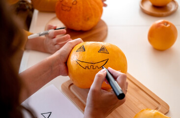 Close up female hands with black marker drawing spooky face on pumpkin. Halloween party preparations.