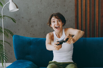 Young fun woman wear white tank shirt play pc game with joystick console sit on blue sofa couch stay home flat rest relax spend free spare time in living room indoors grey wall People lounge concept.