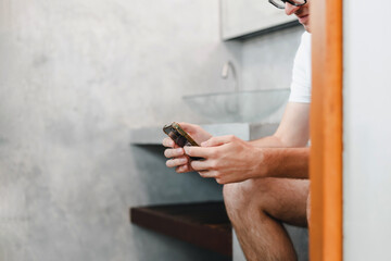 Young Caucasian man gamer sitting on toilet bowl in restroom playing game online on mobile smartphone gadget. Addiction dependency habit social network media technology. Man using phone.
