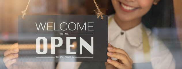 beautiful female business retail owner in medical face mask hanging open wooden sign board at the entrance door of the shop and ready to service customer.