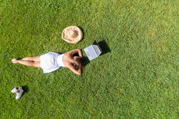 Woman in a white dress, hat with laptop, lying on the green grass at summer day. Top view, drone, aerial view