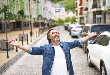 Giving thanks to God mature man standing outdoors with hands spread wide open wearing jeans shirt with white t-shirt under. Mid Aged man glad that life is beautiful standing in old town street