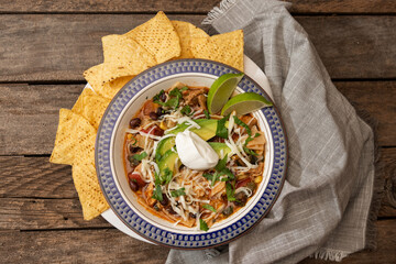 Chicken tortilla soup with sour cream, avocado, lime, tortilla chips and napkin on wooden table, from above up close