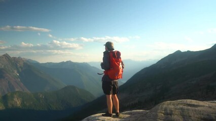caucasian man on hiking trip walks out to cliff edge to view epic mountain landscape