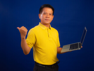 Portrait of smiling middle-aged asian man in casual yellow polo shirt holding laptop isolated on blue background in studio, looking happiness and attractive professional leadership