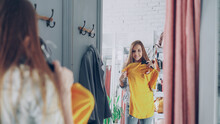 Mirror Shot Of Young Lady Choosing Clothes In Fitting Room. Girl Is Trying Top Checking Size And Length While Standing Opposite Large Mirror. Nice Clothing Store In Background.