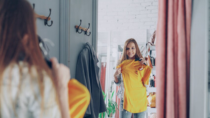 Mirror shot of young lady choosing clothes in fitting room. Girl is trying top checking size and length while standing opposite large mirror. Nice clothing store in background.